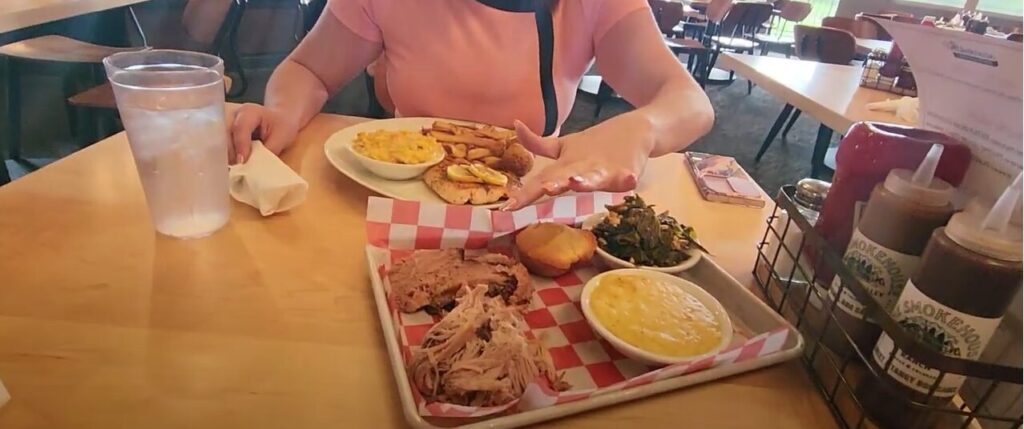 A plate of grilled salmon with lemon, hush puppies, french fries, and creamed corn, alongside a plate of brisket, pulled pork, cheesy grits, collard greens, and cornbread